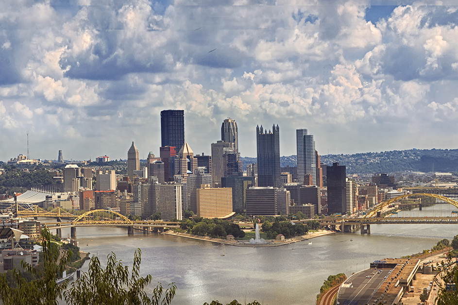 Pittsburgh, PA cityscape with the point in front showing two rivers converging to one with several yellow steel bridges and blue skies with white clouds.