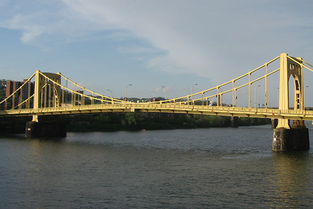 Yellow steel bridge with stone pillars over river with blue and cloudy sky.