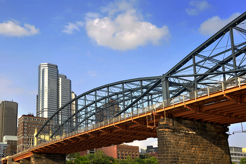 Yellow and black steel bridge with stone pillar with buildings in background with blue sky and white clouds.