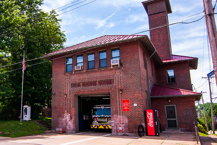 Red brick firehouse building with firetruck in garage and coca cola vending machine outside with tree and American flag.