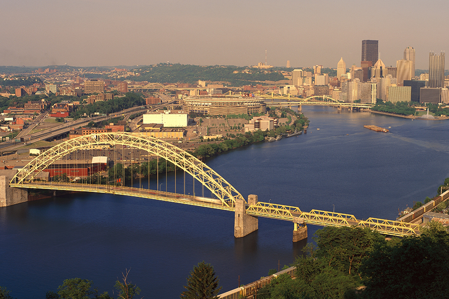 Pittsburgh cityscape with yellow steel bridge in front over blue river.