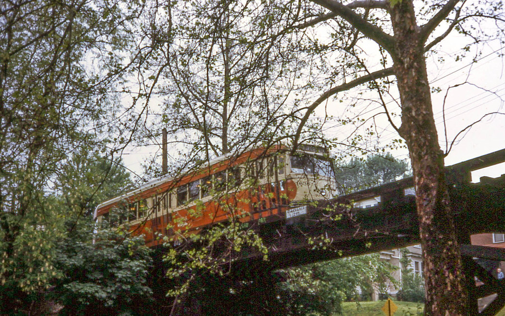 Orange and cream colored train traveling over bridge behind trees.