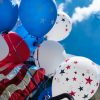 red white and blue balloons with red white and blue stars on the balloons along with an american flag with a bright blue sky in the background